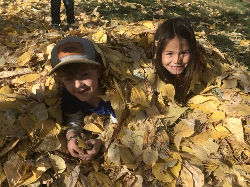 Two children joyfully buried in a pile of autumn leaves at Pine Creek Nature School
